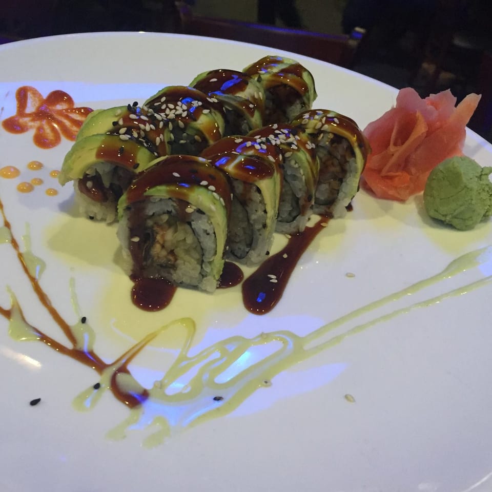 Dragon roll sushi with avocado, eel sauce, sesame seeds, ginger, and wasabi on a white plate.